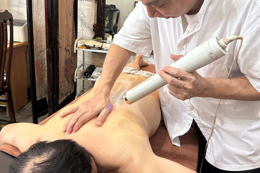 Người thực hiện dùng dụng cụ trị liệu trên vùng lưng của khách nằm sấp, tay còn lại giữ vùng cơ