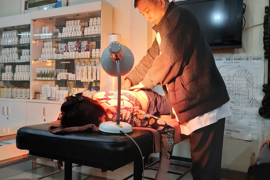 Người thực hiện bấm huyệt vùng lưng cho khách nằm trên bàn trị liệu, bên cạnh là đèn hồng ngoại đang bật