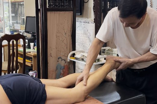 Lương y Đinh Công Lục dùng hai tay tác động vùng bắp chân cho khách nằm sấp trên bàn trị liệu, phía sau tường treo tranh huyệt đạo